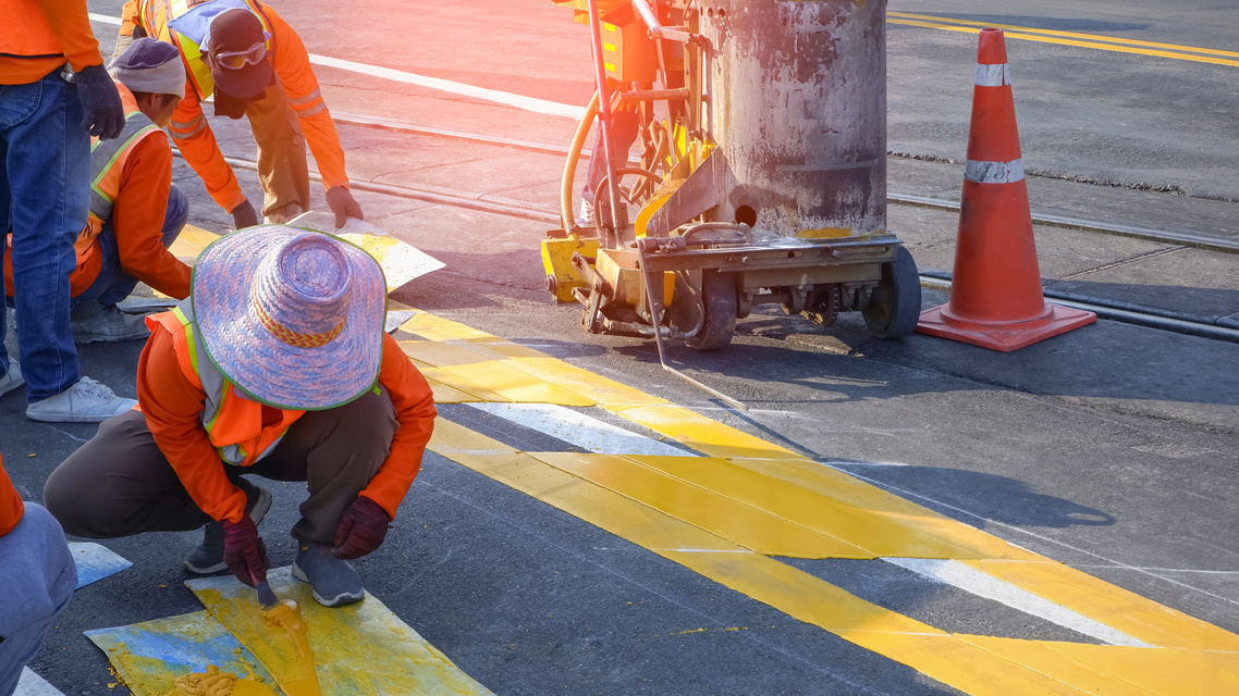 Road workers in reflective vests with thermoplastic spray road marking machine are working to paint traffic yellow lines on asphalt road with railway track crossing on street surface